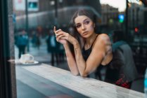 Shot through glass of young brunette posing at counter in cafeteria and looking out away — Stock Photo