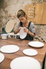 Mujer concentrada en delantal sentada a la mesa y formando platos de arcilla blanca . - foto de stock