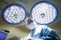 Vue du bas du chirurgien en uniforme debout sous des lampes lumineuses dans la salle d'opération et regardant loin . — Photo de stock