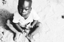 Yoff, Senegal - 6 de diciembre de 2017: Retrato de bajo ángulo del niño sentado en la arena y mirando a la cámara - foto de stock