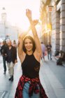 Portrait of stylish brunette holding hands up and cheerfully posing at street in sunlight. — Stock Photo
