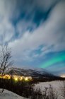 Distant view of illuminated small northern village under cloudy sky in winter night. — Stock Photo