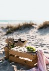 Натюрморт из бокалов с вином и тарелки с белым виноградом, стоящим на деревянной коробке на пляже . — стоковое фото