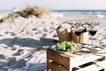 Two glasses of red wine and plate with white grape on timber box on sandy beach. — Stock Photo