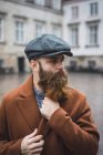 Retrato del hombre barbudo con abrigo y gorra posando en la escena callejera - foto de stock