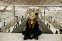 Retrato de mulher bonita de chapéu de malha amarelo sentado no chão na passagem do metrô e olhando para a câmera — Fotografia de Stock