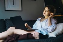 Junge sinnliche Frau liegt mit Laptop auf Sofa und setzt Kopfhörer auf — Stockfoto