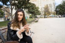Porträt einer brünetten Frau, die mit Smartphone auf einer Parkbank sitzt und wegschaut — Stockfoto