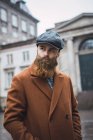 Portrait of bearded man wearing vintage coat and cap posing at square and looking away — Stock Photo