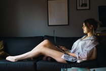 Vue latérale de la femme en chemise couchée avec ordinateur portable sur canapé — Photo de stock