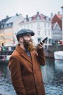 Side view of man in coat and cap using voice search on smartphone at river in city — Stock Photo