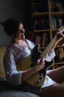 Sorridente giovane donna seduta e suonare la chitarra elettrica a casa . — Foto stock