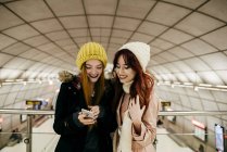 Retrato de duas jovens mulheres de pé e navegando smartphone no shopping . — Fotografia de Stock