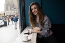Retrato de morena jovem sentada à mesa do café com café e smartphone e olhando para a câmera — Fotografia de Stock
