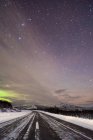 Perspectiva del camino de asfalto cubierto de nieve bajo el cielo estrellado con luces boreales - foto de stock