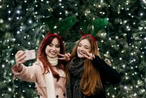 Усміхнені жінки беруть селфі на смартфон на ялинку з освітленням — стокове фото
