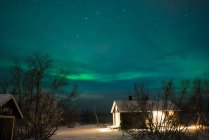 Vista esterna di un piccolo edificio in legno nella foresta invernale sullo sfondo delle luci del nord in cielo — Foto stock