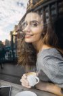 Vue à travers la fenêtre de la femme souriante avec tasse de café regardant la caméra — Photo de stock