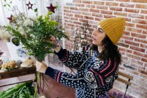 Frau mit Hut und Strickpullover hält Blumenstrauß in der Hand und arrangiert Blumen, während sie im Blumenatelier steht. — Stockfoto