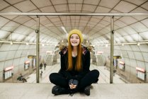 Menina sorridente em chapéu amarelo sentado no chão na passagem do metrô e olhando para a câmera — Fotografia de Stock
