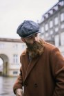 Elegante uomo barbuto in posa in cappotto e cappuccio vintage e guardando oltre spalla — Foto stock