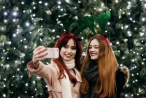 Cheerful pretty women taking selfie with smartphone at festive Christmas tree on street. — Stock Photo