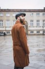Side view of bearded man in retro coat and cap posing at city scene — Stock Photo