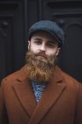 Retrato del hombre barbudo con abrigo marrón y gorra mirando a la cámara - foto de stock