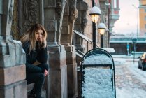 Portrait of bored blonde woman sitting on facade at winter street and looking at camera — Stock Photo