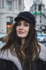 Portrait of young brunette woman in stylish cap looking at camera at street scene — Stock Photo