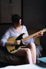 Portrait of woman in shirt sitting on coach and playing guitar — Stock Photo
