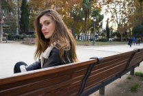 Porträt einer brünetten Frau, die im Park auf einer Bank sitzt und über die Schulter in die Kamera schaut — Stockfoto