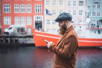 Vue latérale de l'homme élégant naviguant smartphone sur bateau amarré à jetée de la ville sur fond — Photo de stock