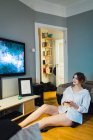 Женщина сидит со смартфоном на полу и смотрит на телевизор дисплей на стене дома — стоковое фото