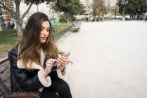 Young pretty woman sitting on bench and using smartphone in urban park. — Stock Photo