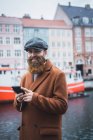 Портрет усміхненого чоловіка, який переглядає смартфон на річці в місті і дивиться на камеру — стокове фото