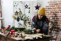 Портрет флориста в трикотажном свитере и шляпе, делающего рождественский венок в цветочном ателье — стоковое фото