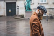 Вид сбоку стильного бородатого мужчины, позирующего в кепке и пальто на городской сцене — стоковое фото