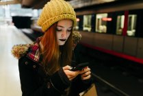 Portrait of young girl in knitted hat using smartphone at subway station — Stock Photo
