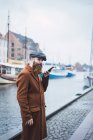Side view of bearded man in coat and cap using voice search on smartphone at river in city — Stock Photo
