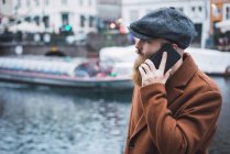 Side view of bearded man in cap talking on smartphone at river in city — Stock Photo