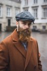 Front view of bearded man wearing vintage coat and cap posing at city scene and looking at camera — Stock Photo