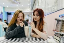 Zwei hübsche junge Frauen sitzen im Café und trinken zusammen einen Cocktail. — Stockfoto