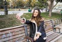 Donna sorridente scattare selfie con smartphone sulla panchina del parco — Foto stock