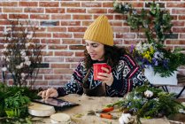 Retrato de la mujer bebiendo café y y y usando tableta en la mesa en atelier floral - foto de stock