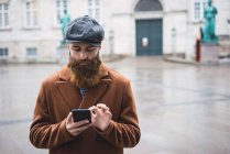 Homem barbudo de casaco e boné usando smartphone na rua da cidade . — Fotografia de Stock