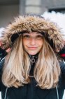 Retrato de mulher loira sorridente no capuz com peles mulher posando na cidade de inverno e olhando para a câmera — Fotografia de Stock