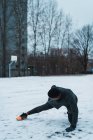 Vista lateral del hombre atlético en cuclillas en el suelo cubierto de nieve y la pierna extendida para calentar los músculos - foto de stock