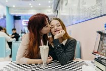 Portrait of redhead girl kissing girlfriends cheek at cafe table with milk shake — Stock Photo