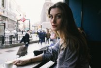 Junge brünette Frau schaut in die Kamera, während sie im Café sitzt und Kaffee trinkt. — Stockfoto